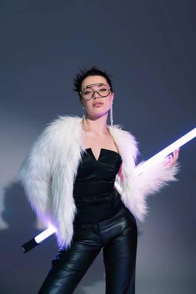 Trendy woman in black corset and white faux fur jacket posing with fluorescent lamp on grey background — Stock Photo