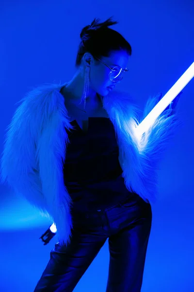 Young woman in black corset and furry jacket posing with fluorescent lamp on bright blue background — Stock Photo