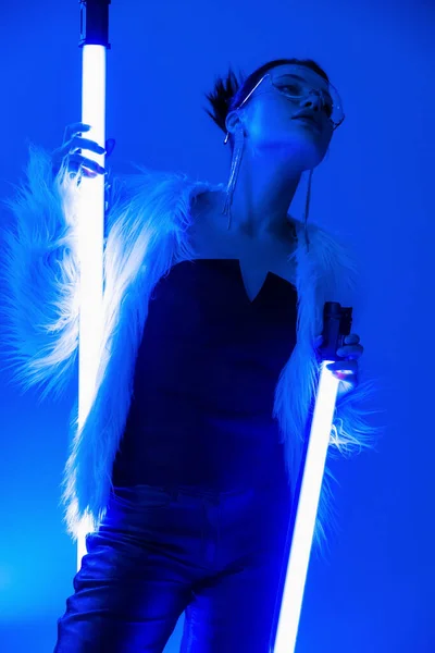 Low angle view of trendy woman in faux fur jacket and black corset holding fluorescent lamps on blue background — Stock Photo