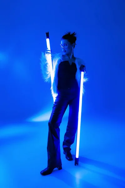 Полная длина стильная женщина в белом искусственном меховом пиджаке держа лампы на синем фоне — стоковое фото