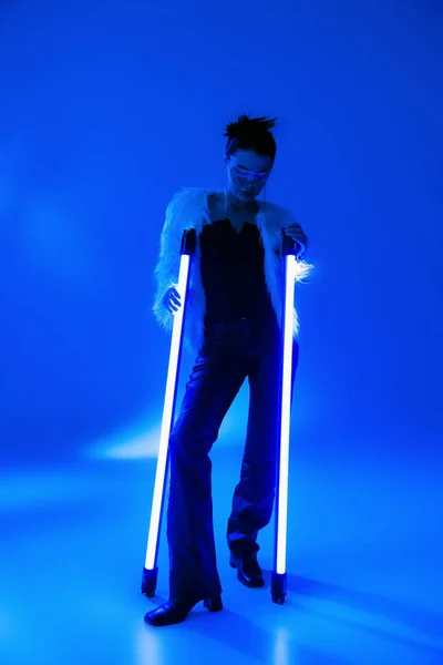 Pleine longueur de femme à la mode en fausse fourrure veste tenant des lampes sur fond bleu — Photo de stock