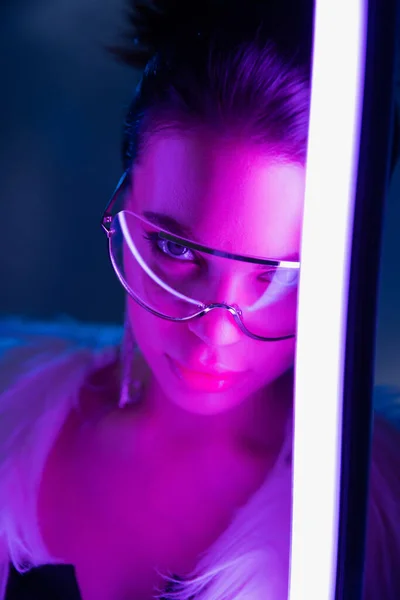 Trendy junge Frau mit Sonnenbrille blickt in die Kamera in der Nähe Lampe auf marineblauem Hintergrund — Stockfoto