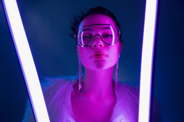 Mujer joven de moda en gafas de sol y chaqueta de piel sintética mirando a la cámara cerca de lámparas en fondo azul marino - foto de stock