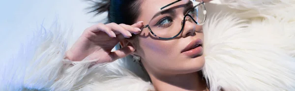 Vista dall'alto della donna elegante in giacca di pelliccia sintetica che tocca gli occhiali da sole su sfondo bianco con illuminazione blu, banner — Foto stock