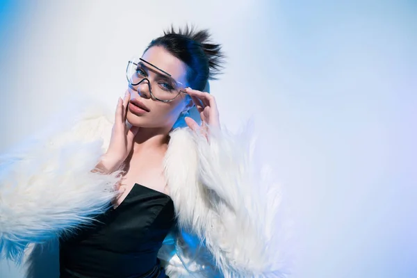 Вид зверху на стильну жінку в штучній шубці і сонцезахисних окулярах, дивлячись на камеру на білому тлі з синім освітленням — стокове фото