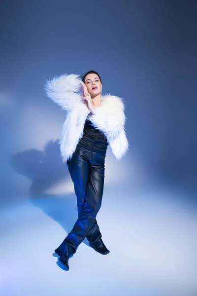 Full length of trendy brunette model in faux fur jacket posing on blue background — Stock Photo