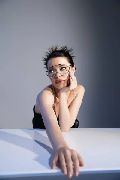 Jeune femme branchée en lunettes de soleil regardant loin près de la table sur fond gris — Photo de stock