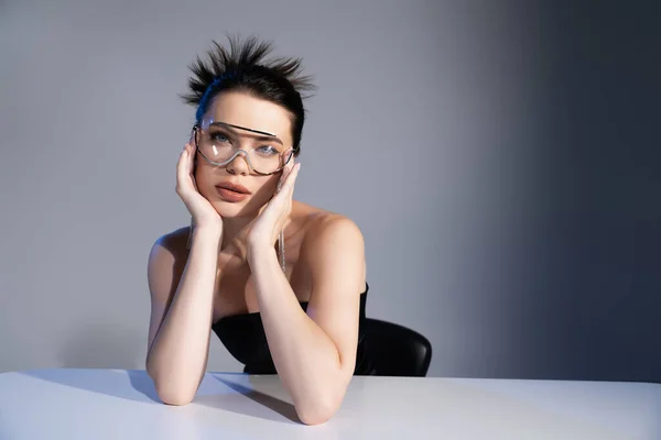 Jolie jeune modèle en lunettes de soleil regardant la caméra près de la table sur fond gris — Photo de stock