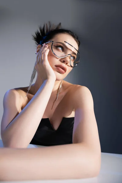 Modisches Modell in Korsett und Sonnenbrille schaut am Tisch vor grauem Hintergrund weg — Stockfoto