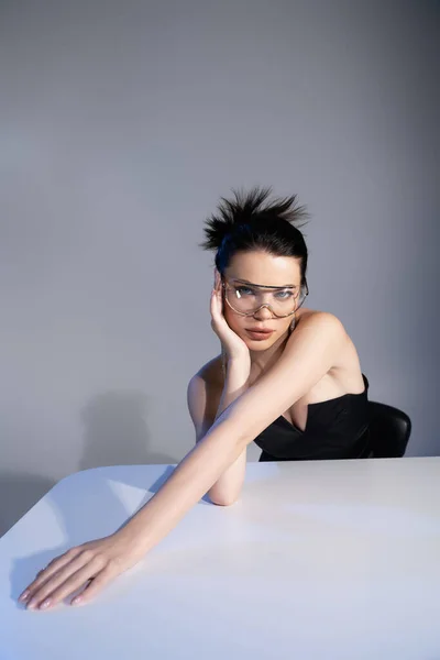 Hübsche brünette Frau mit Sonnenbrille berührt Gesicht neben Tisch auf grauem Hintergrund — Stockfoto