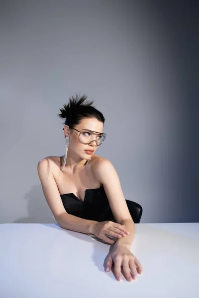 Femme élégante en corset noir et lunettes de soleil posant près de la table sur fond gris — Photo de stock