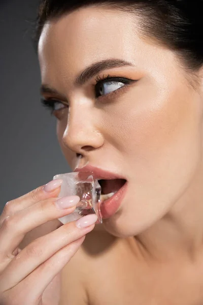 Porträt einer brünetten Frau mit Make-up, die Eiswürfel in der Nähe der Lippen hält, isoliert auf grau — Stockfoto