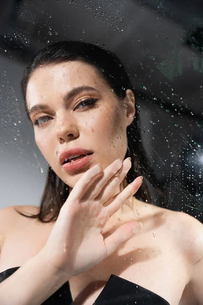 Sexy woman with makeup touching wet glass on grey background — Stock Photo