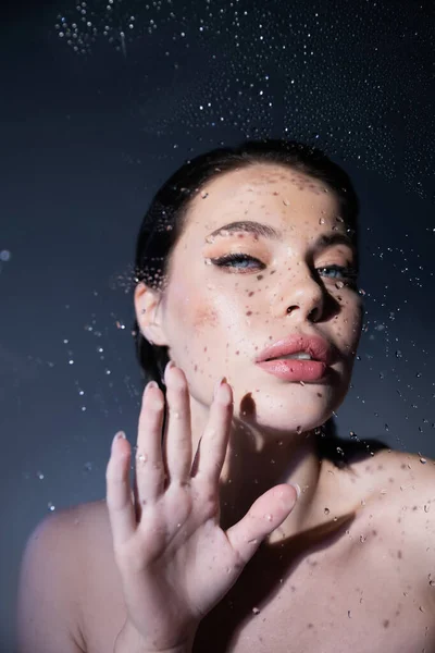 Сексуальная модель с голыми плечами, касающаяся мокрого стекла на сером фоне — стоковое фото
