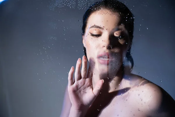 Jovem mulher com ombros nus olhando para o vidro molhado em fundo cinza — Fotografia de Stock