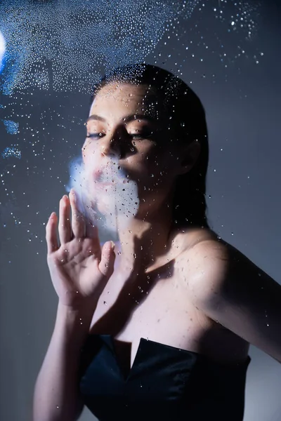 Stylish brunette woman breathing on wet glass on grey background — Stock Photo