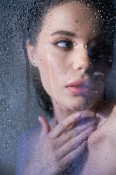 Jeune femme brune touchant le cou derrière du verre mouillé sur fond gris — Photo de stock