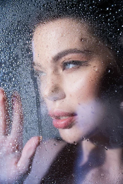 Young woman touching blurred glass with water drops on grey background — Stock Photo