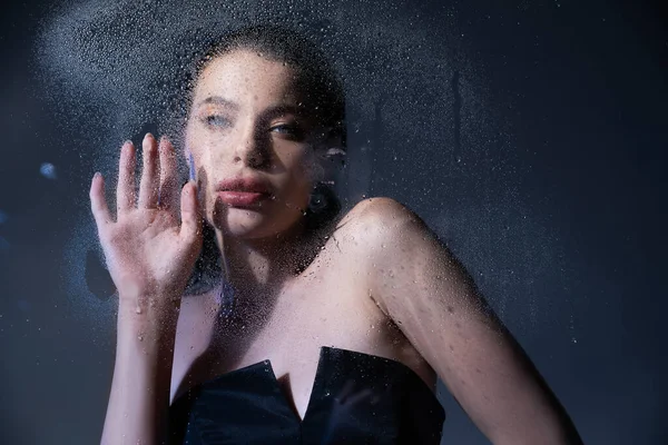 Elegante donna in corsetto toccare vetro bagnato su sfondo grigio — Stock Photo