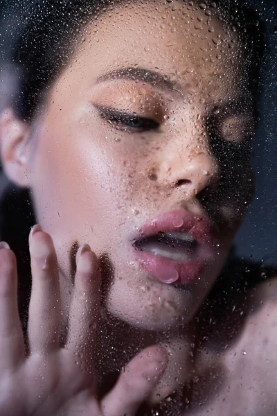 Mujer borrosa con maquillaje tocando vidrio con gotas de agua sobre fondo gris - foto de stock