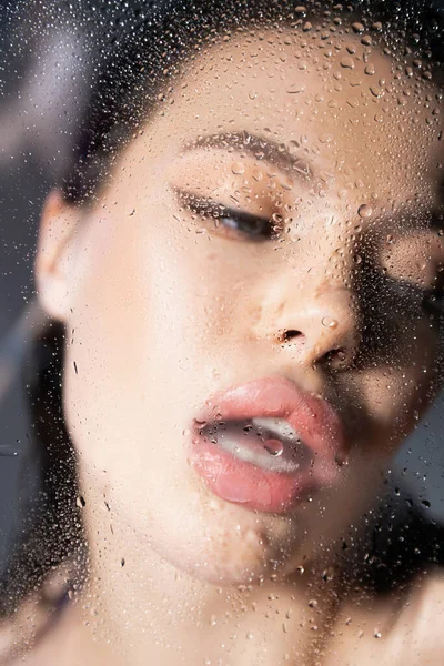 Sexy woman standing behind blurred wet glass on grey background — Stock Photo