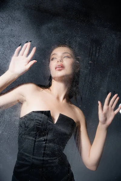 Low angle view of fashionable woman in corset standing behind wet glass on grey background — Stock Photo