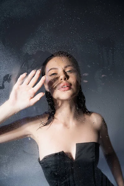 Модна брюнетка модель в корсеті, торкаючись мокрого скла на сірому фоні — стокове фото