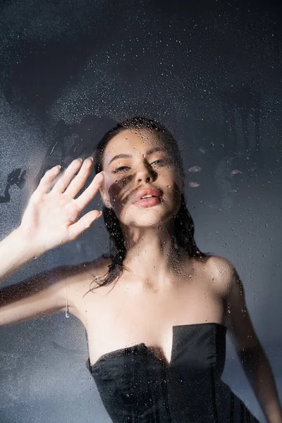 Trendy brunette model in corset touching wet glass on grey background — Stock Photo