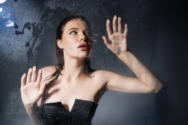 Junge Frau in schwarzem Korsett berührt nasses Glas auf grauem Hintergrund — Stockfoto