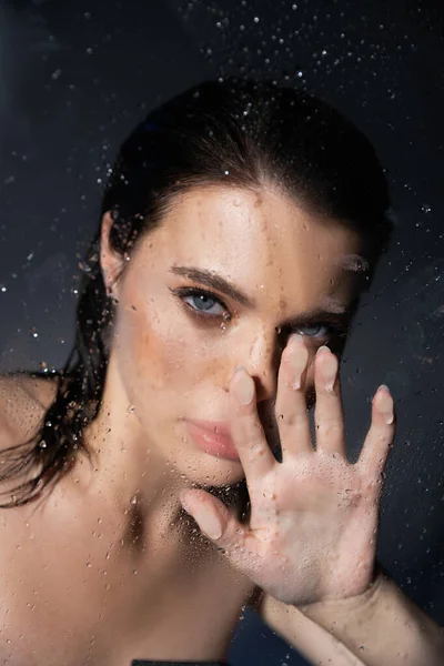 Jeune femme brune avec maquillage touchant verre humide sur fond gris — Photo de stock