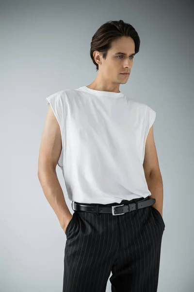 Hombre con estilo en camiseta y pantalones negros de pie con las manos en los bolsillos en gris - foto de stock