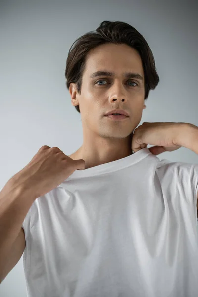Homem elegante olhando para a câmera e ajustando t-shirt branca no cinza — Fotografia de Stock