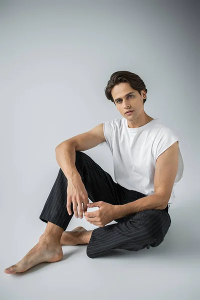 Homme pieds nus en pantalon rayé et t-shirt blanc assis sur gris — Photo de stock