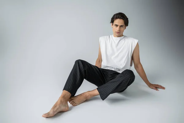 Homme pieds nus en pantalon rayé et t-shirt blanc regardant la caméra tout en étant assis sur gris — Photo de stock