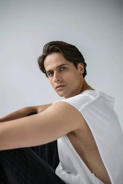 Brunette man in striped trousers and white t-shirt looking at camera while sitting isolated on grey — Stock Photo