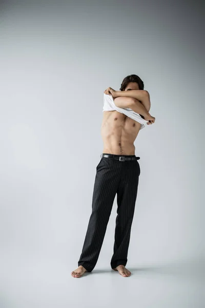 Full length of sexy and muscular man in trousers taking off white t-shirt on grey — стоковое фото