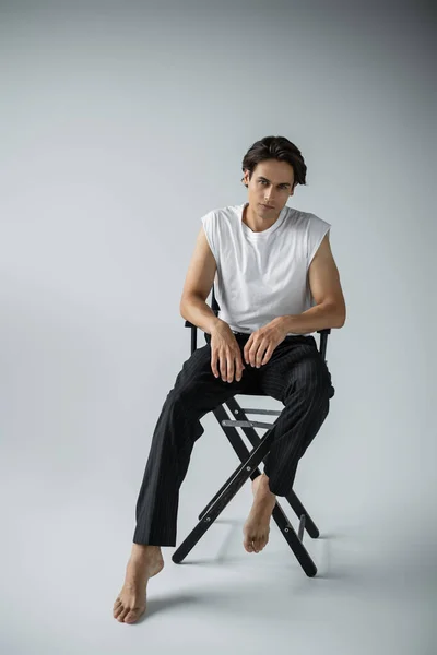 Full length of barefoot man in trousers and white t-shirt sitting on camp chair on grey — Stock Photo