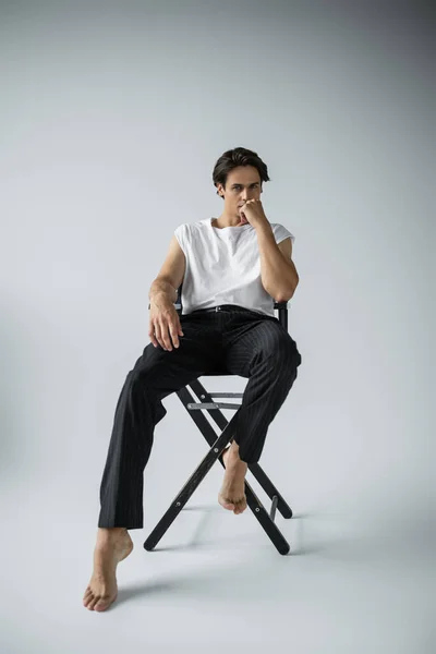 Homme pieds nus en pantalon rayé et t-shirt blanc assis sur chaise de camp sur gris — Photo de stock