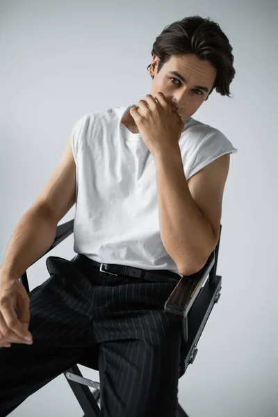 Brunette homme en pantalon rayé et t-shirt blanc assis sur chaise de camp sur gris — Photo de stock