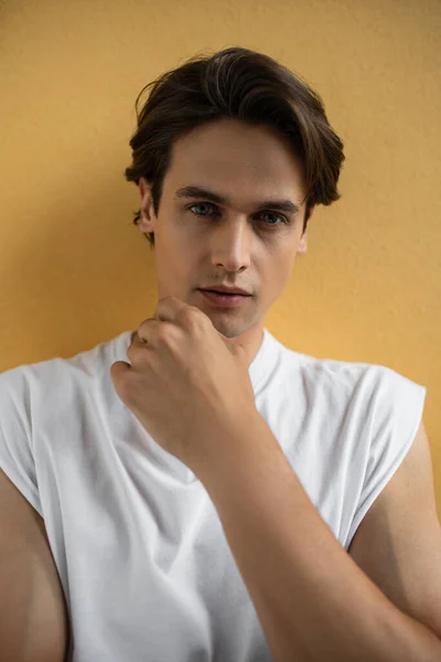 Hombre con estilo en camiseta blanca mirando a la cámara mientras está de pie cerca de la pared amarilla - foto de stock