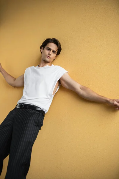 Hombre con estilo en camiseta blanca y pantalones a rayas posando mientras mira la cámara cerca de la pared amarilla - foto de stock