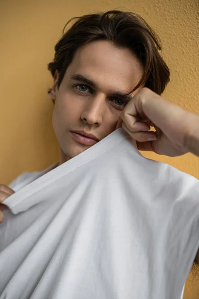 Portrait of brunette man adjusting white t-shirt and posing near yellow wall — Stock Photo