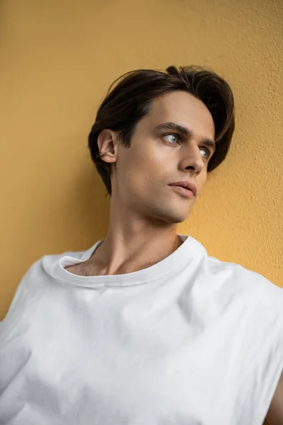 Hombre con estilo en camiseta blanca mirando hacia otro lado mientras posa cerca de la pared amarilla - foto de stock