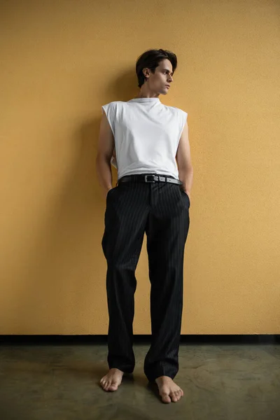 Longitud completa de hombre con estilo en camiseta blanca y pantalones a rayas posando con las manos en bolsillos cerca de la pared amarilla - foto de stock