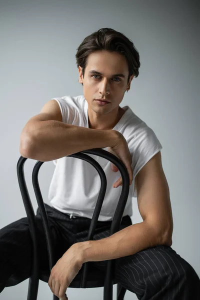 Brunette man in white t-shirt and pants leaning on chair while posing on grey — Stock Photo