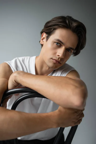 Morena hombre en blanco camiseta apoyada en silla mientras posando en gris - foto de stock