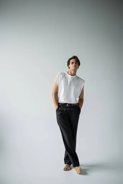 Homme élégant en t-shirt blanc et pantalon noir posant debout avec les jambes croisées sur le gris — Photo de stock
