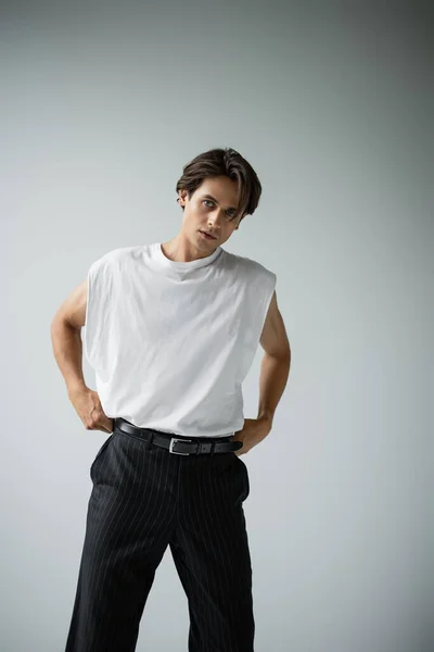 Hombre con estilo en camiseta blanca y pantalones negros posando mientras mira a la cámara en gris - foto de stock