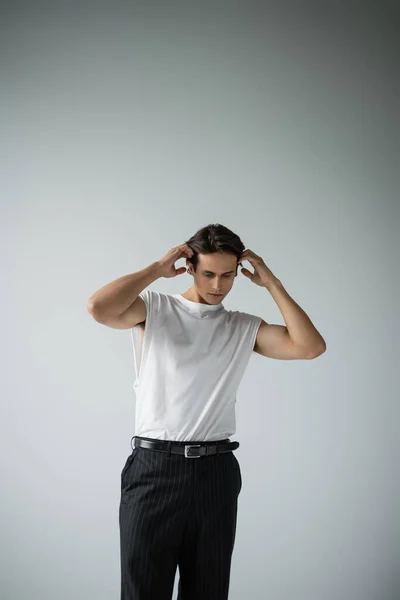 Stylish man in white t-shirt and black pants posing while adjusting hair on grey — Stock Photo