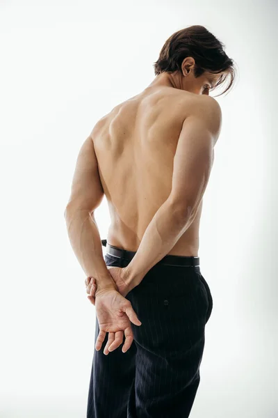 Back view of muscular and shirtless man in black trousers posing with hands behind back on white — Stock Photo